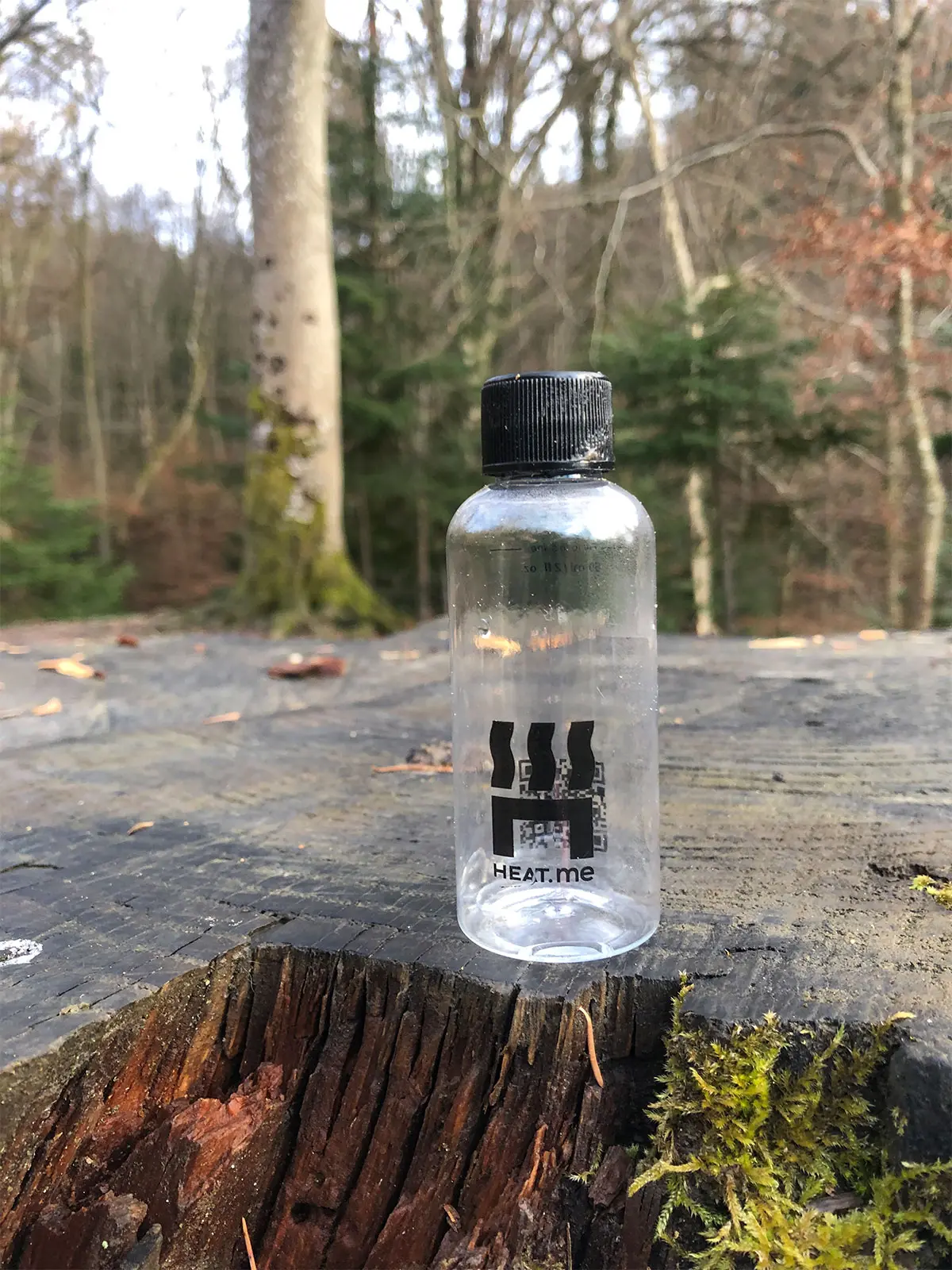 Dosierflasche auf einem Baumstumpf im Wald. In diese Flasche passen bis zur Markierung 60 ml Wasser. Diese Menge an Wasser wird benötigt, um mit dem HEAT.me Heizpad Essen zu erhitzen.