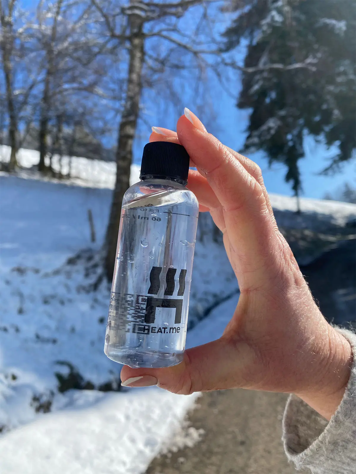Dosierflasche zwischen zwei Fingern in Schneelandschaft. Die Dosierflasche ist für 60 ml Wasser gedacht, um unterwegs mit dem HEAT.me Lunchbox Kit ein heißes Essen oder Getränk zu machen.