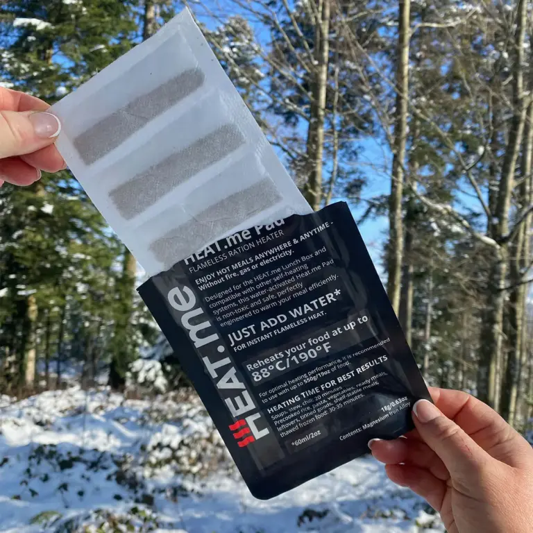 In einer Winterlandschaft hält die eine Hand die Verpackung vom HEAT.me Heizpad - einem Flameless Ration Heater (FRH) und die andere Hand zieht das Heizpad aus der Verpackung.