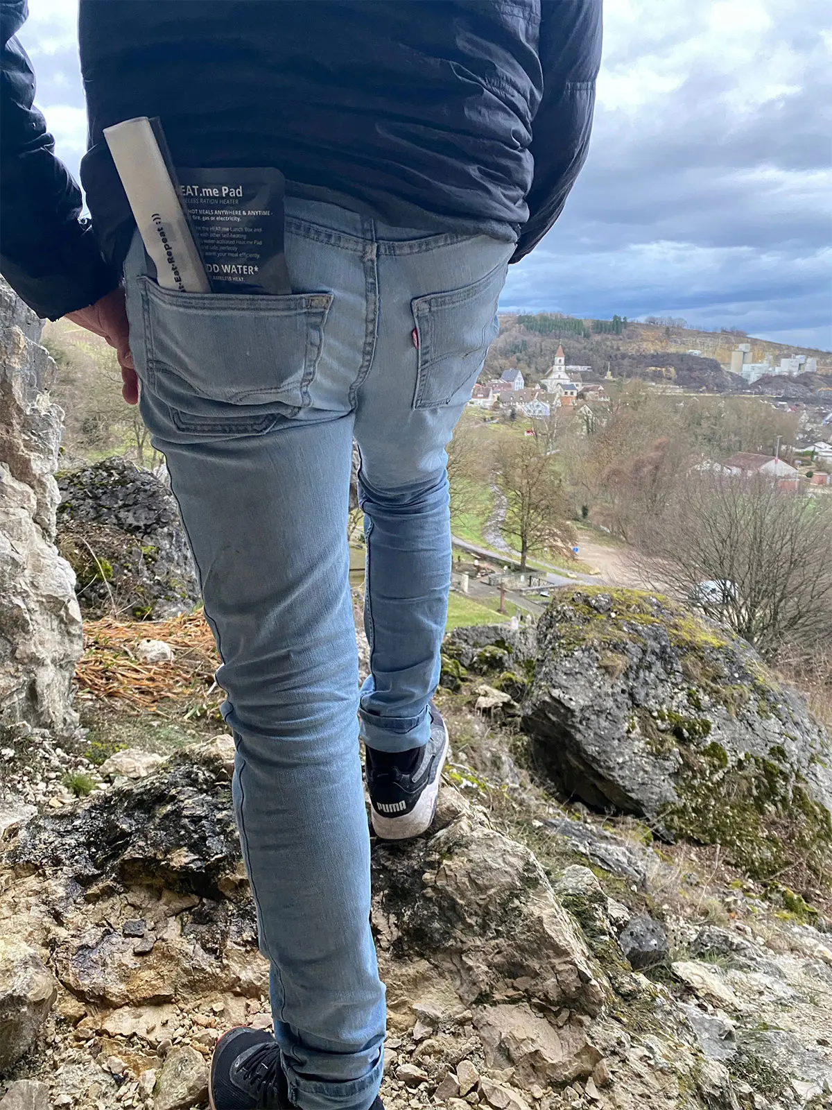 Mann mit blauen Jeans, der eine zusammengerollte HEAT.me Lunchbag und ein HEAT.me Heizpad in der hinteren Hosentasche hat, auf einem Felsen stehend