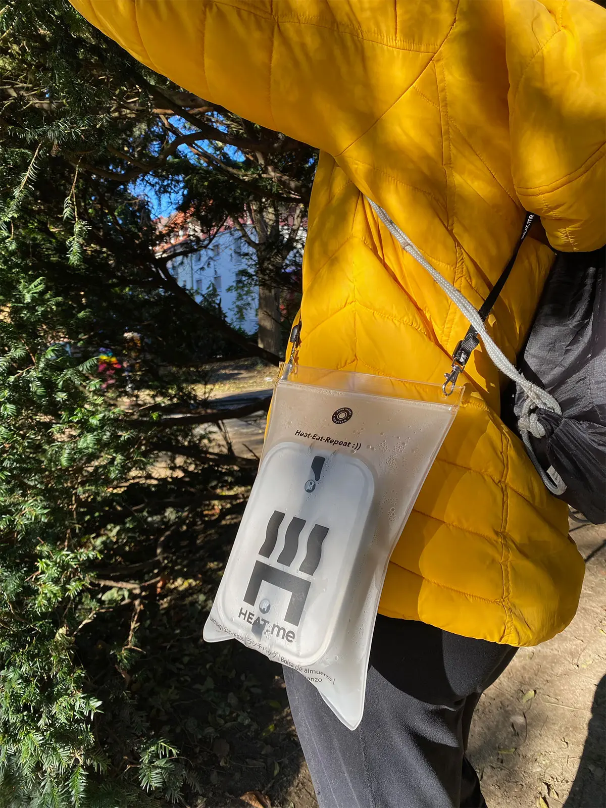 Frau mit gelber Jacke trägt ein HEAT.me Lunchbox-Kit mit beheizbarer Lunchbox an der Schulter, bereit für ein warmes Mittagessen im Freien
