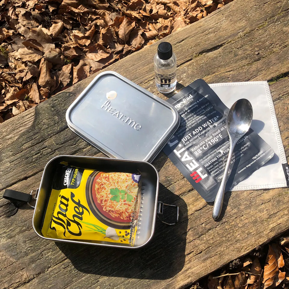 Auf einem Holztisch im Wald steht eine Lunchbox mit Tha-Instantsuppe, daneben der Deckel, die Dosierflasche, das Heizpad, die Schutzhülle und ein Löffel.